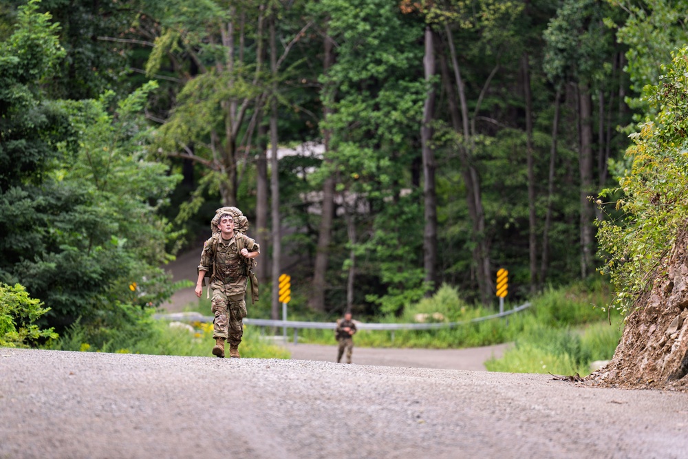 A soldier in full military camouflage uniform and gear is running along a paved road through a forested area, carrying a large rucksack on their back. A second soldier is visible further back on the road, also in uniform. The setting appears to be a military training exercise or ruck march event in a wooded, rural environment.