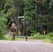 A soldier in full military camouflage uniform and gear is running along a paved road through a forested area, carrying a large rucksack on their back. A second soldier is visible further back on the road, also in uniform. The setting appears to be a military training exercise or ruck march event in a wooded, rural environment.