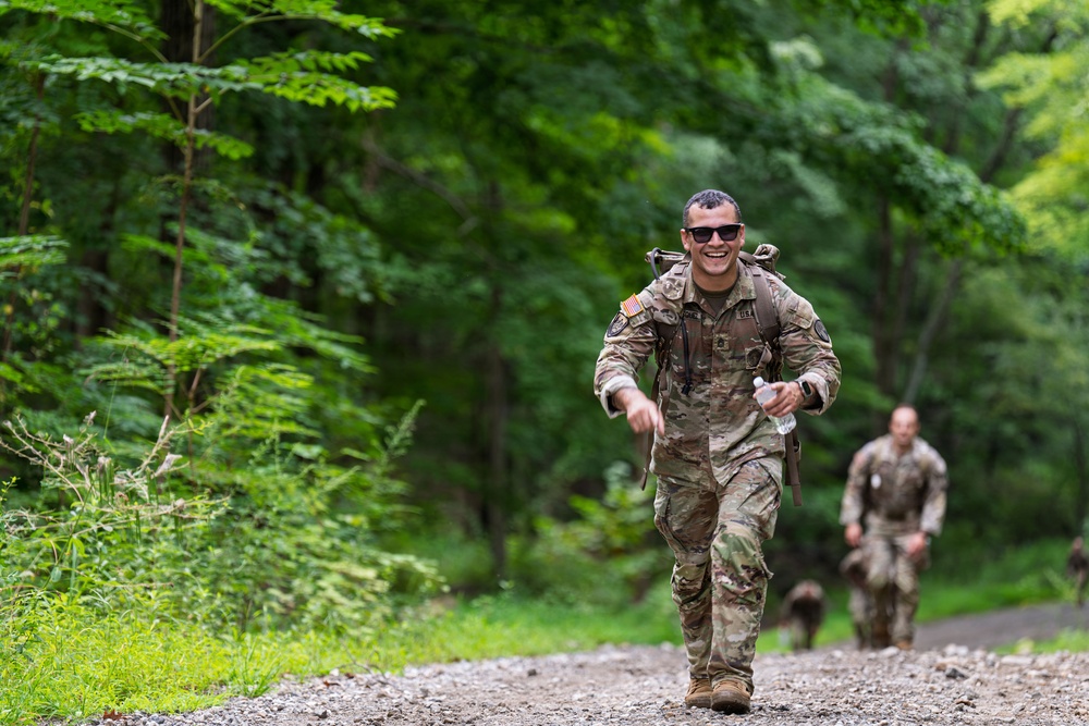 A smiling U.S. Army soldier in full camouflage uniform and rucksack runs along a gravel trail through a dense green forest. He is leading ahead of at least one other soldier visible in the background, suggesting a ruck march or physical training event. The atmosphere appears competitive yet spirited, indicated by the lead soldier's broad smile.