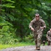 A smiling U.S. Army soldier in full camouflage uniform and rucksack runs along a gravel trail through a dense green forest. He is leading ahead of at least one other soldier visible in the background, suggesting a ruck march or physical training event. The atmosphere appears competitive yet spirited, indicated by the lead soldier's broad smile.