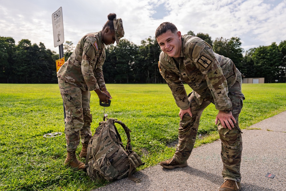 Two U.S. Army soldiers in OCP (Operational Camouflage Pattern) uniforms are standing at the end of a 2-mile run course marker on a military installation. One soldier is smiling toward the camera while leaning forward with hands on knees, appearing fatigued but in good spirits, while the other soldier is handling what appears to be a scale or timing device near a tactical backpack. The setting is an outdoor grassy field area with trees in the background.