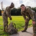Two U.S. Army soldiers in OCP (Operational Camouflage Pattern) uniforms are standing at the end of a 2-mile run course marker on a military installation. One soldier is smiling toward the camera while leaning forward with hands on knees, appearing fatigued but in good spirits, while the other soldier is handling what appears to be a scale or timing device near a tactical backpack. The setting is an outdoor grassy field area with trees in the background.
