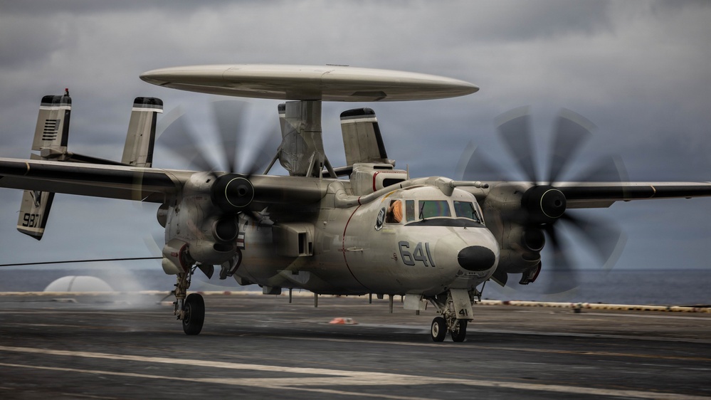 A U.S. Navy E-2 Hawkeye airborne early warning aircraft, marked with the number '641', is taxiing or preparing for launch on the deck of an aircraft carrier. The aircraft's twin turboprop engines are running at high power, evidenced by the motion-blurred propellers. The scene is set under overcast skies with the ocean visible in the background.
