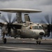 A U.S. Navy E-2 Hawkeye airborne early warning aircraft, marked with the number '641', is taxiing or preparing for launch on the deck of an aircraft carrier. The aircraft's twin turboprop engines are running at high power, evidenced by the motion-blurred propellers. The scene is set under overcast skies with the ocean visible in the background.