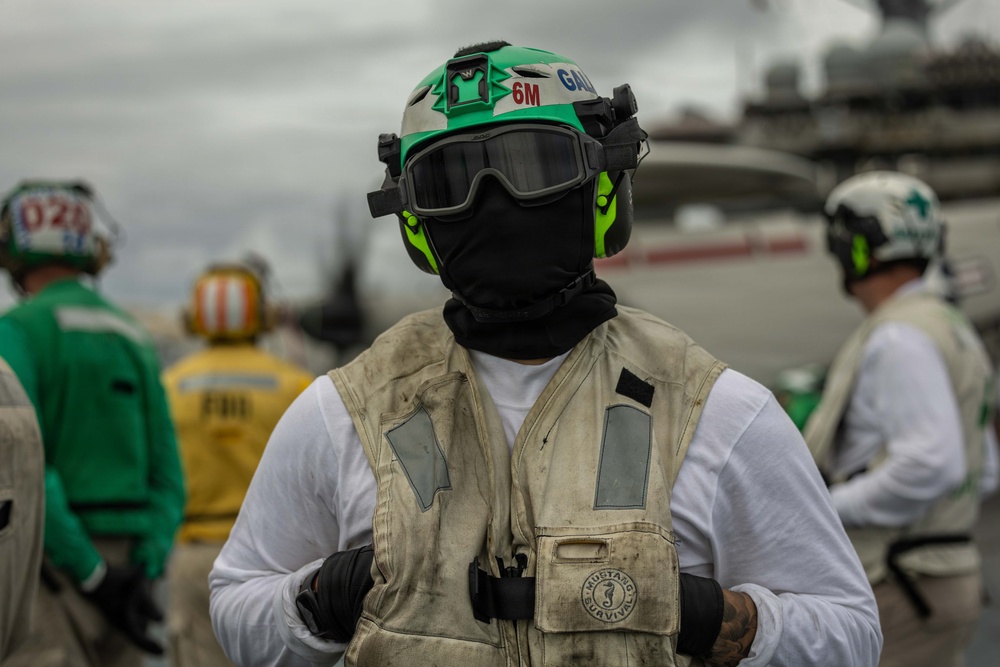 A naval aviation deck crew member stands in the foreground on what appears to be an aircraft carrier flight deck. The individual is wearing standard flight deck safety gear including a green helmet marked '6M' and 'GAL', protective goggles, a black balaclava, and a worn Mustang Survival flotation vest. Several other crew members in green and yellow jerseys are visible in the blurred background.