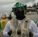 A naval aviation deck crew member stands in the foreground on what appears to be an aircraft carrier flight deck. The individual is wearing standard flight deck safety gear including a green helmet marked '6M' and 'GAL', protective goggles, a black balaclava, and a worn Mustang Survival flotation vest. Several other crew members in green and yellow jerseys are visible in the blurred background.