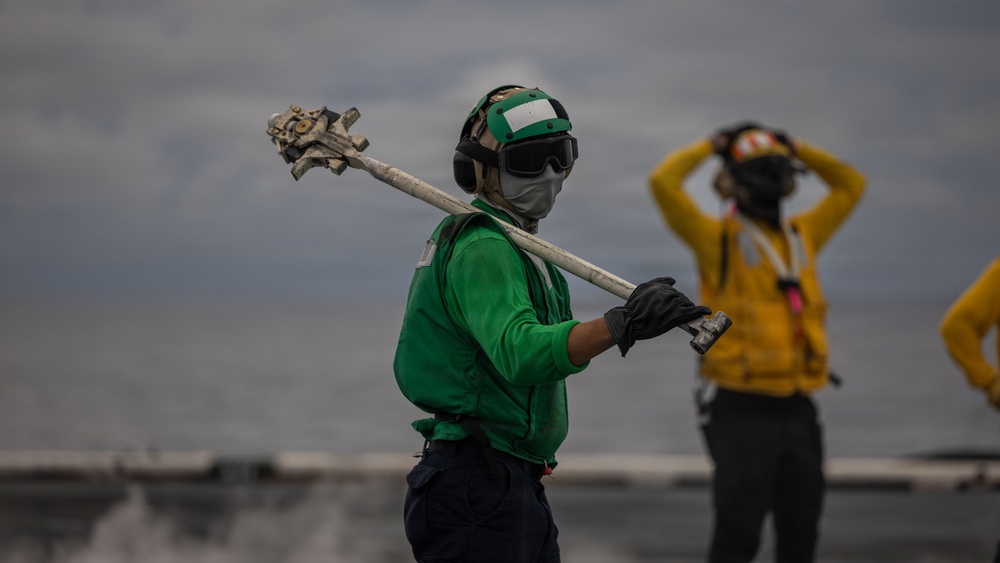 The image shows naval aviation deck crew members working on what appears to be an aircraft carrier flight deck. The foreground figure wears a green jersey, helmet, goggles, and face mask while carrying a long-handled tool over their shoulder. A second crew member in a yellow jersey is visible in the background with hands raised to their helmet.