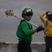 The image shows naval aviation deck crew members working on what appears to be an aircraft carrier flight deck. The foreground figure wears a green jersey, helmet, goggles, and face mask while carrying a long-handled tool over their shoulder. A second crew member in a yellow jersey is visible in the background with hands raised to their helmet.