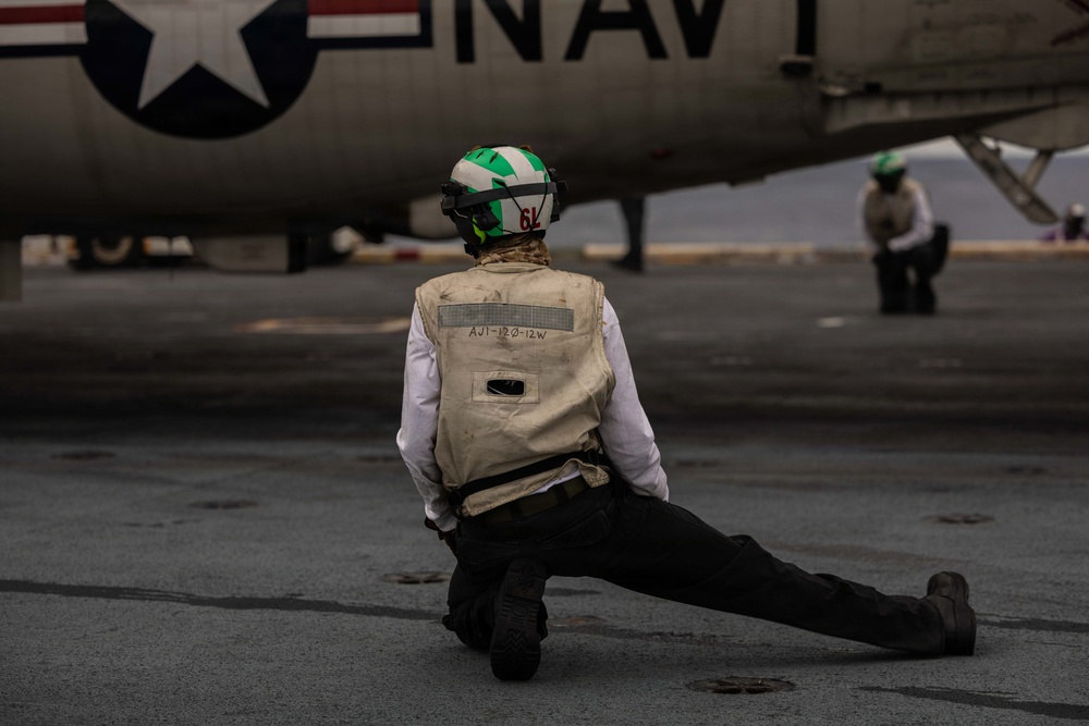 A U.S. Navy deck crew member is kneeling on an aircraft carrier flight deck, facing a naval aircraft marked with 'NAVY' and a U.S. roundel insignia. The crew member wears a green-and-white helmet numbered '6L' and a tan vest marked 'AJ1-120-12W', consistent with carrier flight deck operations. A second crew member is visible in the background near another aircraft.