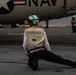 A U.S. Navy deck crew member is kneeling on an aircraft carrier flight deck, facing a naval aircraft marked with 'NAVY' and a U.S. roundel insignia. The crew member wears a green-and-white helmet numbered '6L' and a tan vest marked 'AJ1-120-12W', consistent with carrier flight deck operations. A second crew member is visible in the background near another aircraft.