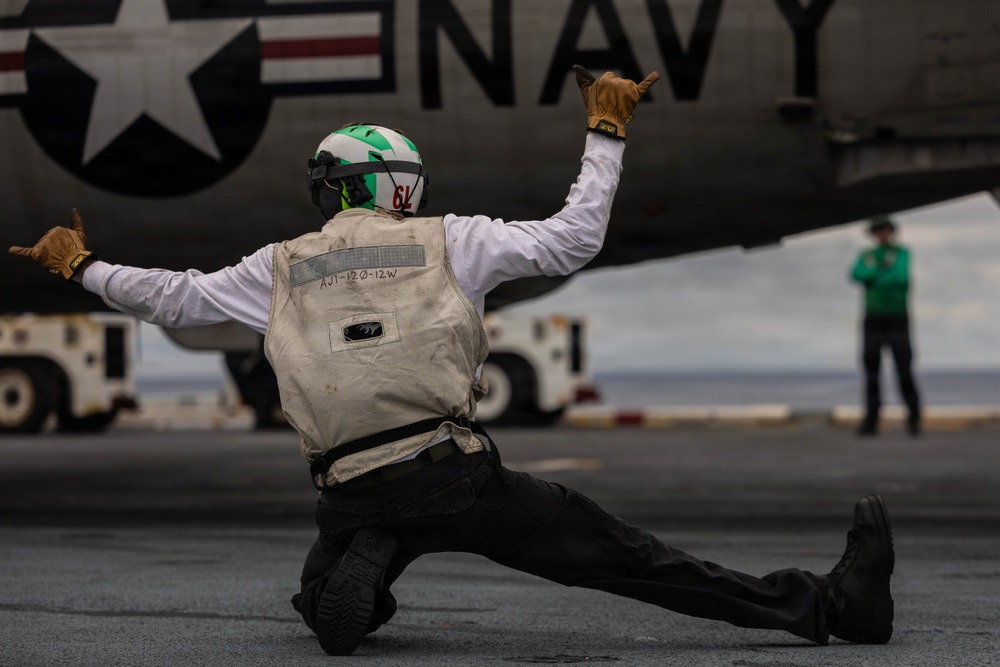 A U.S. Navy flight deck crew member is captured in a dynamic crouching pose with arms outstretched, signaling on an aircraft carrier flight deck. The individual is positioned directly beneath a Navy aircraft displaying the U.S. roundel insignia and 'NAVY' lettering. A second crew member in green is visible in the background near deck equipment.