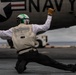 A U.S. Navy flight deck crew member is captured in a dynamic crouching pose with arms outstretched, signaling on an aircraft carrier flight deck. The individual is positioned directly beneath a Navy aircraft displaying the U.S. roundel insignia and 'NAVY' lettering. A second crew member in green is visible in the background near deck equipment.