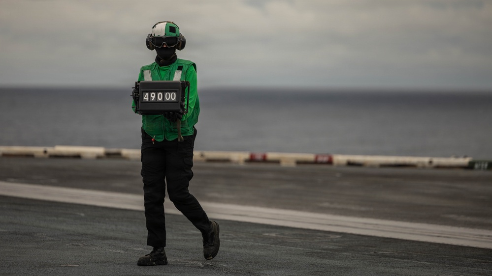 A naval aviation deck crew member wearing a green jersey and helmet with face covering stands on an aircraft carrier flight deck holding a weight board displaying '49000'. The green jersey identifies this individual as a catapult and arresting gear officer or aircraft handling crew member. The ocean horizon is visible in the background under overcast skies.