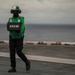 A naval aviation deck crew member wearing a green jersey and helmet with face covering stands on an aircraft carrier flight deck holding a weight board displaying '49000'. The green jersey identifies this individual as a catapult and arresting gear officer or aircraft handling crew member. The ocean horizon is visible in the background under overcast skies.