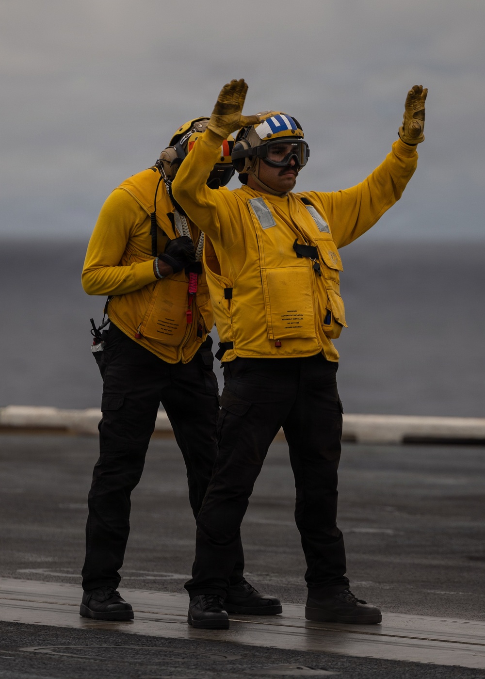 Two naval personnel in yellow flight deck jerseys and safety gear are performing hand signals on what appears to be a wet aircraft carrier flight deck. The foreground figure is giving a raised-arm signal while wearing a helmet with blue and white markings, goggles, and a yellow float coat. Overcast skies and a visible ocean horizon confirm an at-sea operational environment.