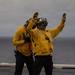 Two naval personnel in yellow flight deck jerseys and safety gear are performing hand signals on what appears to be a wet aircraft carrier flight deck. The foreground figure is giving a raised-arm signal while wearing a helmet with blue and white markings, goggles, and a yellow float coat. Overcast skies and a visible ocean horizon confirm an at-sea operational environment.