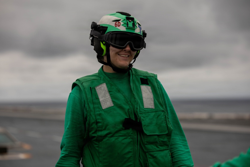 A person wearing green flight deck gear stands on what appears to be an aircraft carrier flight deck. They are smiling and equipped with a helmet marked '6D', protective goggles, ear protection, and a green float coat with reflective patches. The overcast sky and visible ocean horizon confirm an at-sea naval environment.