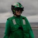 A person wearing green flight deck gear stands on what appears to be an aircraft carrier flight deck. They are smiling and equipped with a helmet marked '6D', protective goggles, ear protection, and a green float coat with reflective patches. The overcast sky and visible ocean horizon confirm an at-sea naval environment.