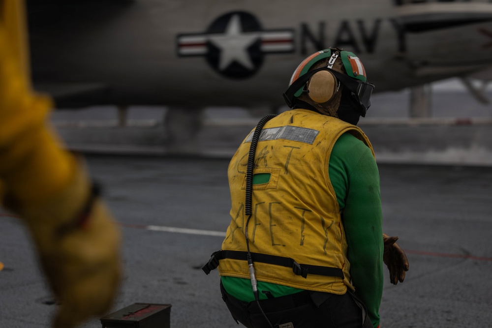 A U.S. Navy flight deck crew member wearing a yellow safety vest and green jersey stands on an aircraft carrier flight deck. The personnel is facing away from the camera, wearing a helmet with ear protection and a visor, with a U.S. Navy aircraft visible in the background. The yellow vest has text reading 'CAT SAFETY' written on the back, indicating this individual is a catapult safety observer.