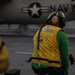 A U.S. Navy flight deck crew member wearing a yellow safety vest and green jersey stands on an aircraft carrier flight deck. The personnel is facing away from the camera, wearing a helmet with ear protection and a visor, with a U.S. Navy aircraft visible in the background. The yellow vest has text reading 'CAT SAFETY' written on the back, indicating this individual is a catapult safety observer.