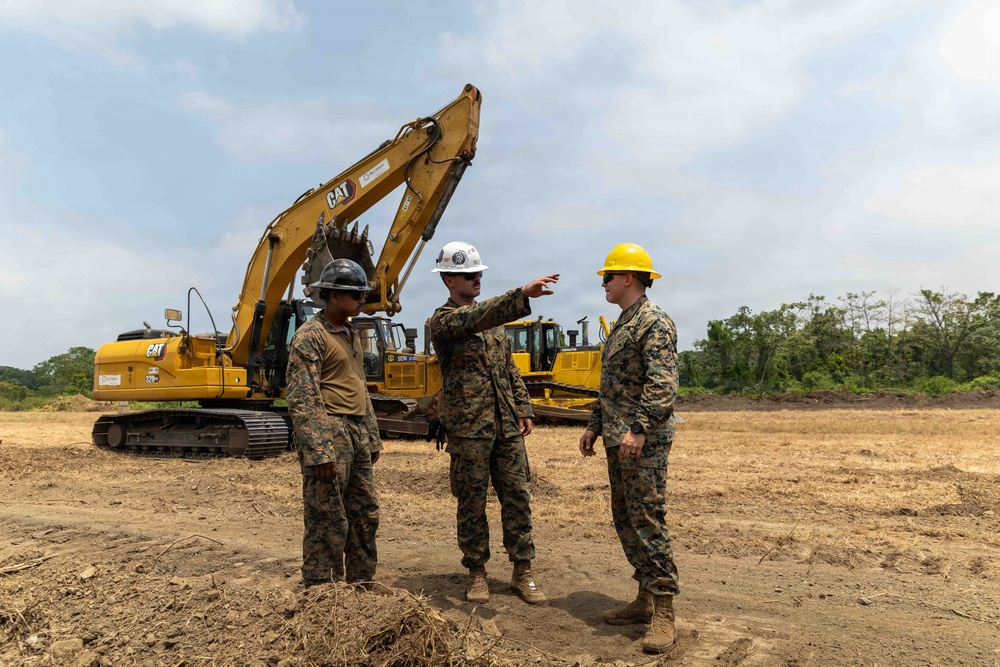 Three individuals in military camouflage uniforms are gathered at an active construction or earthmoving site. One person wearing a white hard hat is pointing in a direction, apparently giving instructions or briefing the others. Large CAT (Caterpillar) heavy construction equipment, including an excavator and what appears to be a bulldozer, are visible in the background.