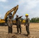 Three individuals in military camouflage uniforms are gathered at an active construction or earthmoving site. One person wearing a white hard hat is pointing in a direction, apparently giving instructions or briefing the others. Large CAT (Caterpillar) heavy construction equipment, including an excavator and what appears to be a bulldozer, are visible in the background.