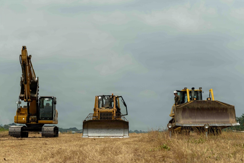 Three large yellow Caterpillar construction machines are positioned across an open, dry grassy field under an overcast sky. The equipment includes one tracked excavator on the left and two bulldozers in the center and right. At least one operator wearing a white hard hat is visible on the rightmost bulldozer.