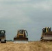 Three large yellow Caterpillar construction machines are positioned across an open, dry grassy field under an overcast sky. The equipment includes one tracked excavator on the left and two bulldozers in the center and right. At least one operator wearing a white hard hat is visible on the rightmost bulldozer.