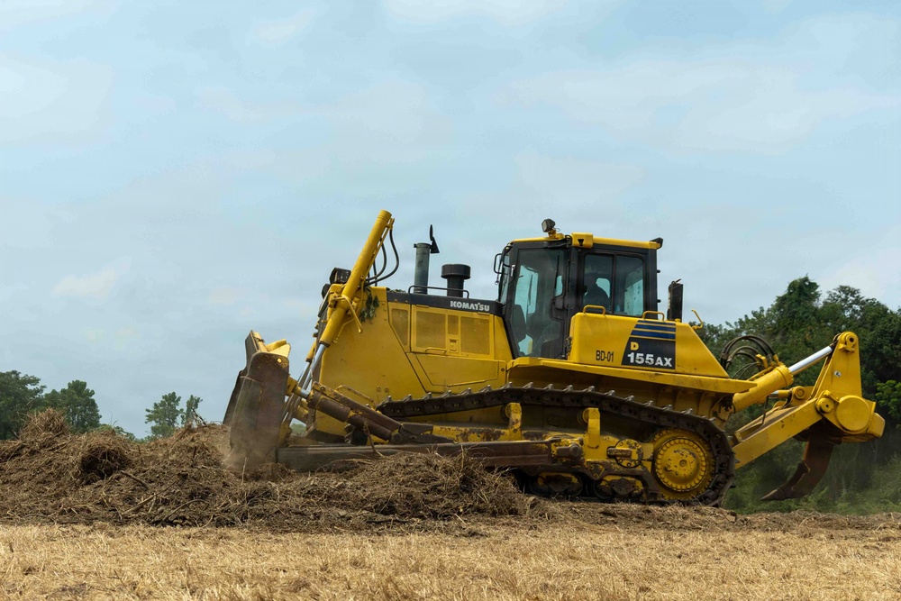 A yellow Komatsu D155AX bulldozer is actively pushing and clearing soil and vegetation debris on an open field. The machine is identified with the unit marking 'BD-01' and appears to be in operation during land clearing or grading work. The setting is an outdoor construction or agricultural site with trees visible in the background under a partly cloudy sky.