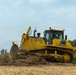 A yellow Komatsu D155AX bulldozer is actively pushing and clearing soil and vegetation debris on an open field. The machine is identified with the unit marking 'BD-01' and appears to be in operation during land clearing or grading work. The setting is an outdoor construction or agricultural site with trees visible in the background under a partly cloudy sky.