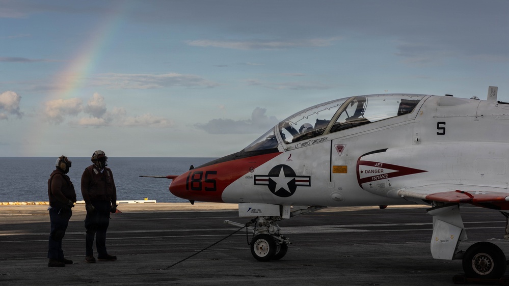 A U.S. Navy T-45 Goshawk trainer aircraft (bureau number 185) is positioned on the flight deck of an aircraft carrier at sea. Two flight deck personnel wearing helmets and flight gear stand near the nose of the aircraft. A rainbow is visible in the background over the ocean horizon.