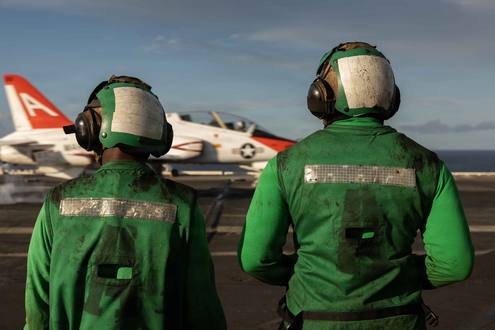 Two U.S. Navy flight deck crew members wearing green jerseys and helmets observe a T-45 Goshawk trainer aircraft on what appears to be an aircraft carrier flight deck. The personnel are positioned with their backs to the camera, watching the red-and-white painted jet. Green jerseys indicate these crew members are likely catapult and arresting gear personnel or aircraft handlers.