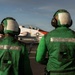 Two U.S. Navy flight deck crew members wearing green jerseys and helmets observe a T-45 Goshawk trainer aircraft on what appears to be an aircraft carrier flight deck. The personnel are positioned with their backs to the camera, watching the red-and-white painted jet. Green jerseys indicate these crew members are likely catapult and arresting gear personnel or aircraft handlers.