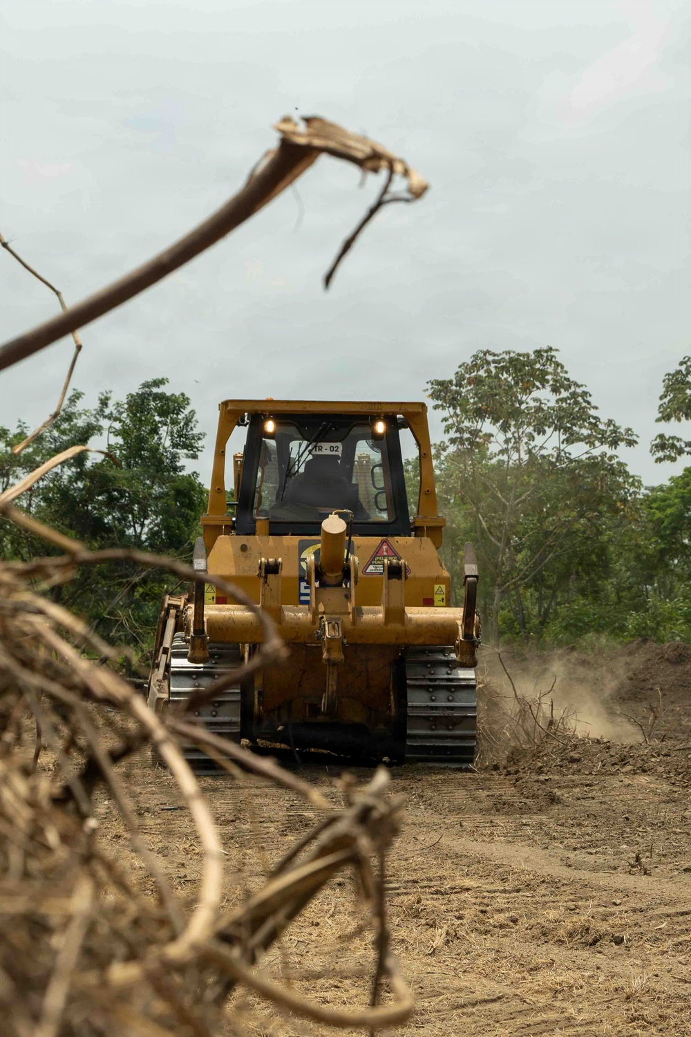 A yellow crawler bulldozer, marked 'IR-02' on its cab, is operating on a cleared dirt path surrounded by felled vegetation and dust. The machine is moving forward through what appears to be a land-clearing or road-construction operation in a tropical or subtropical environment. The foreground contains dry, dead branches framing the scene.