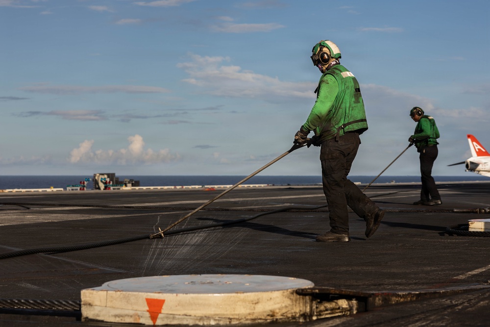 Two personnel in green jerseys and cranial helmets are washing down the flight deck of an aircraft carrier at sea. The foreground worker is using a long-handled squeegee or hose tool to clean the deck surface. A red-tailed aircraft is partially visible in the background on the right side of the deck.