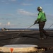 Two personnel in green jerseys and cranial helmets are washing down the flight deck of an aircraft carrier at sea. The foreground worker is using a long-handled squeegee or hose tool to clean the deck surface. A red-tailed aircraft is partially visible in the background on the right side of the deck.