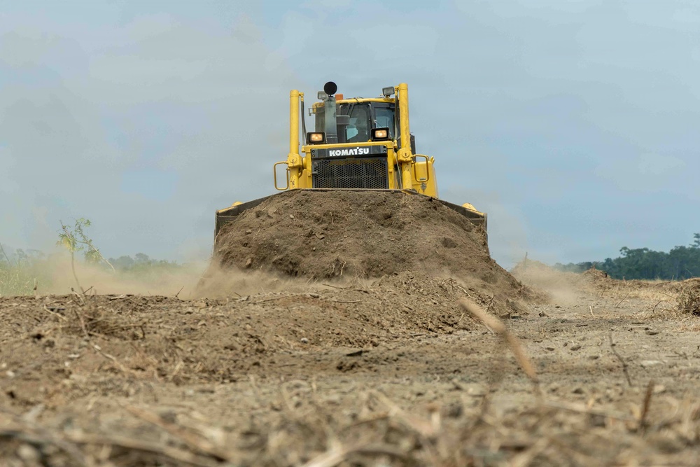 A yellow Komatsu bulldozer is actively pushing a large mound of dry earth across a cleared, barren landscape. The machine is captured head-on, with dust billowing from the sides of the blade as it moves soil. Vegetation and what appears to be forest tree line is visible in the distant background, suggesting land clearing operations.