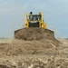A yellow Komatsu bulldozer is actively pushing a large mound of dry earth across a cleared, barren landscape. The machine is captured head-on, with dust billowing from the sides of the blade as it moves soil. Vegetation and what appears to be forest tree line is visible in the distant background, suggesting land clearing operations.