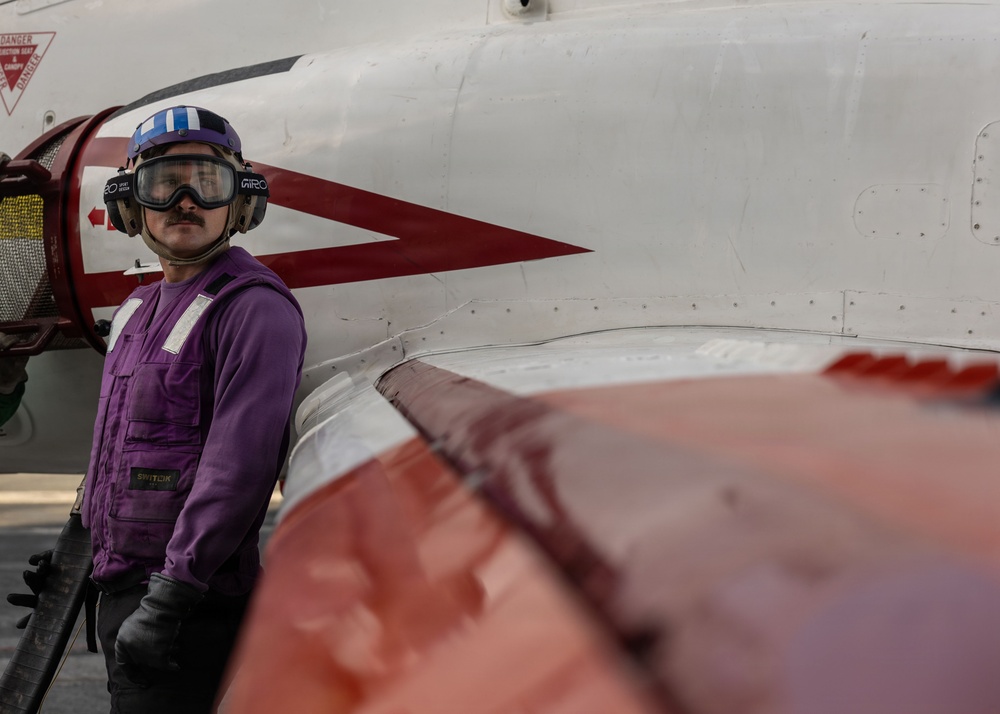 A Navy aviation fueler (identified by purple jersey/vest) stands beside a military aircraft on what appears to be an aircraft carrier flight deck. The personnel is wearing standard flight deck safety gear including a purple helmet with goggles and hearing protection. A red and white painted aircraft fuselage with warning markings is prominently visible in the foreground and background.