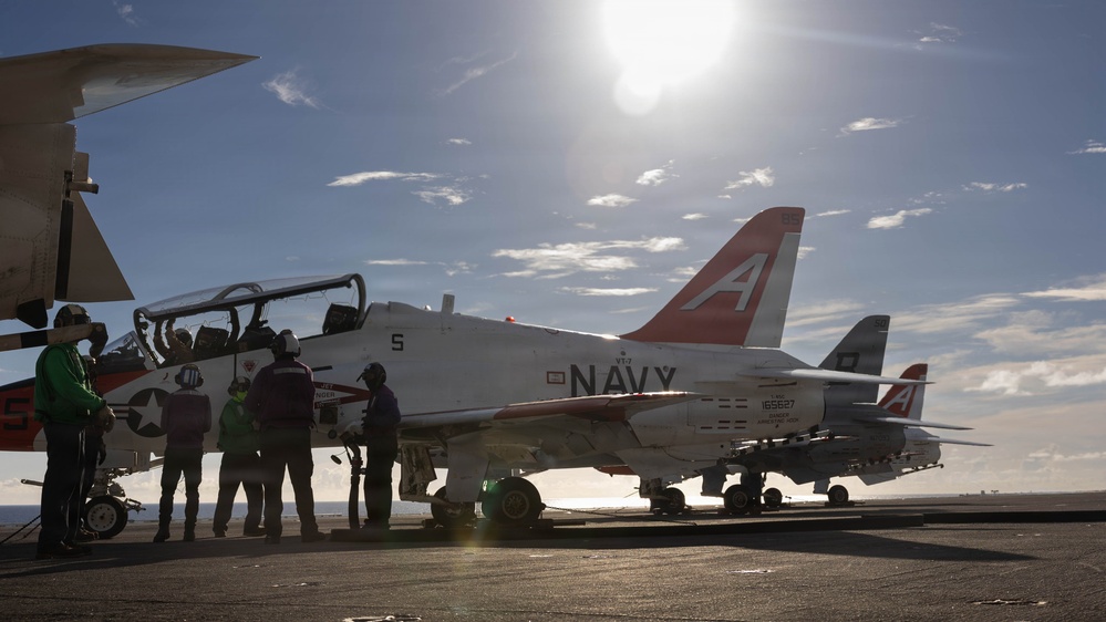 A U.S. Navy T-45 Goshawk trainer aircraft (Bureau Number 165627) is positioned on an aircraft carrier flight deck, marked with squadron designation VT-7 and a distinctive red-and-white tail with the letter 'A'. Several flight deck personnel in colored jerseys are gathered around the aircraft, likely conducting pre-flight checks or maintenance. The image is taken against a bright sun, creating a backlit, high-contrast scene.