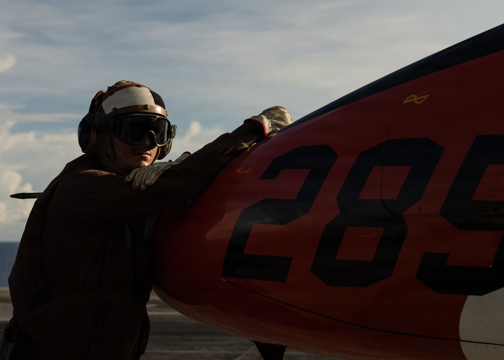 A military deck crew member wearing a flight deck helmet, goggles, and gloves is handling or inspecting a red aircraft component marked with the number '285' on what appears to be an aircraft carrier flight deck. The image is taken in dramatic side lighting, creating strong contrast between the subject and the bright sky background. The setting suggests active flight deck operations at sea.