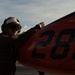 A military deck crew member wearing a flight deck helmet, goggles, and gloves is handling or inspecting a red aircraft component marked with the number '285' on what appears to be an aircraft carrier flight deck. The image is taken in dramatic side lighting, creating strong contrast between the subject and the bright sky background. The setting suggests active flight deck operations at sea.