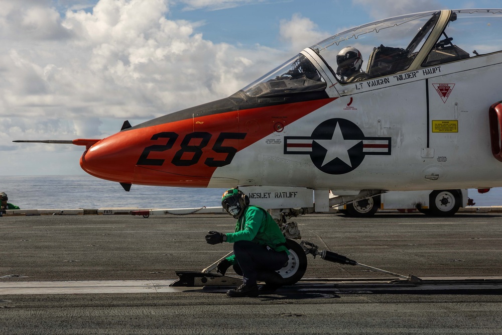 A U.S. Navy T-45 Goshawk training jet (bureau number 285) is positioned on an aircraft carrier flight deck, apparently being prepared for a catapult launch. A deck crew member in a green jersey is crouched near the nose gear, likely inspecting or attaching the catapult launch bar. The aircraft bears the name markings 'LT VAUGHN WILDER HAUPT' and 'WESLEY RATLIFF' on its fuselage.