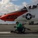 A U.S. Navy T-45 Goshawk training jet (bureau number 285) is positioned on an aircraft carrier flight deck, apparently being prepared for a catapult launch. A deck crew member in a green jersey is crouched near the nose gear, likely inspecting or attaching the catapult launch bar. The aircraft bears the name markings 'LT VAUGHN WILDER HAUPT' and 'WESLEY RATLIFF' on its fuselage.