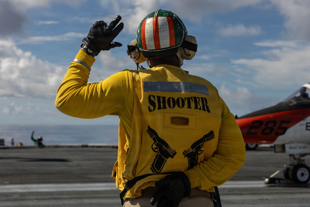 A naval flight deck crew member designated as 'Shooter' stands on an aircraft carrier flight deck, giving a hand signal. The individual is wearing standard yellow flight deck jersey with the role designation 'SHOOTER' printed on the back, along with a tri-color helmet (green, white, red) and hearing protection. An orange training aircraft marked '285' is visible in the background.