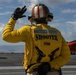 A naval flight deck crew member designated as 'Shooter' stands on an aircraft carrier flight deck, giving a hand signal. The individual is wearing standard yellow flight deck jersey with the role designation 'SHOOTER' printed on the back, along with a tri-color helmet (green, white, red) and hearing protection. An orange training aircraft marked '285' is visible in the background.