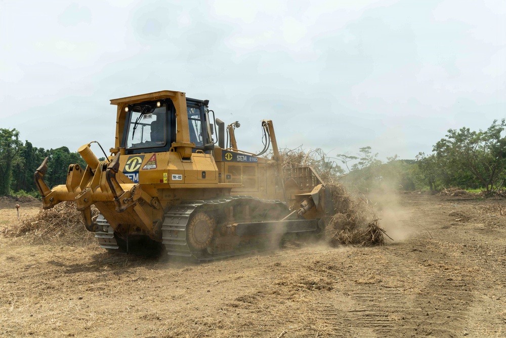A yellow SEM 816D bulldozer is actively clearing land, pushing debris, vegetation, and dry brush across a dusty, barren terrain. The machine appears to be engaged in land preparation or deforestation activity, with visible dust clouds rising from the ground. Trees and green vegetation are visible in the background, suggesting the cleared area borders a forested zone.