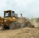 A yellow SEM 816D bulldozer is actively clearing land, pushing debris, vegetation, and dry brush across a dusty, barren terrain. The machine appears to be engaged in land preparation or deforestation activity, with visible dust clouds rising from the ground. Trees and green vegetation are visible in the background, suggesting the cleared area borders a forested zone.