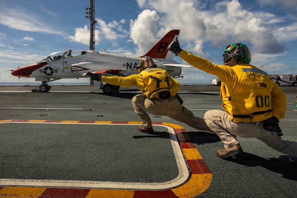 Two naval flight deck crew members in yellow 'SHOOTER' jerseys are signaling the launch of a T-45 Goshawk trainer aircraft on what appears to be a US Navy aircraft carrier flight deck. The aircraft, marked with 'NAV' and the number 285, features the distinctive red and white training color scheme. The shooters are in their characteristic crouching launch-signal pose, pointing toward the catapult direction.