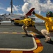 Two naval flight deck crew members in yellow 'SHOOTER' jerseys are signaling the launch of a T-45 Goshawk trainer aircraft on what appears to be a US Navy aircraft carrier flight deck. The aircraft, marked with 'NAV' and the number 285, features the distinctive red and white training color scheme. The shooters are in their characteristic crouching launch-signal pose, pointing toward the catapult direction.
